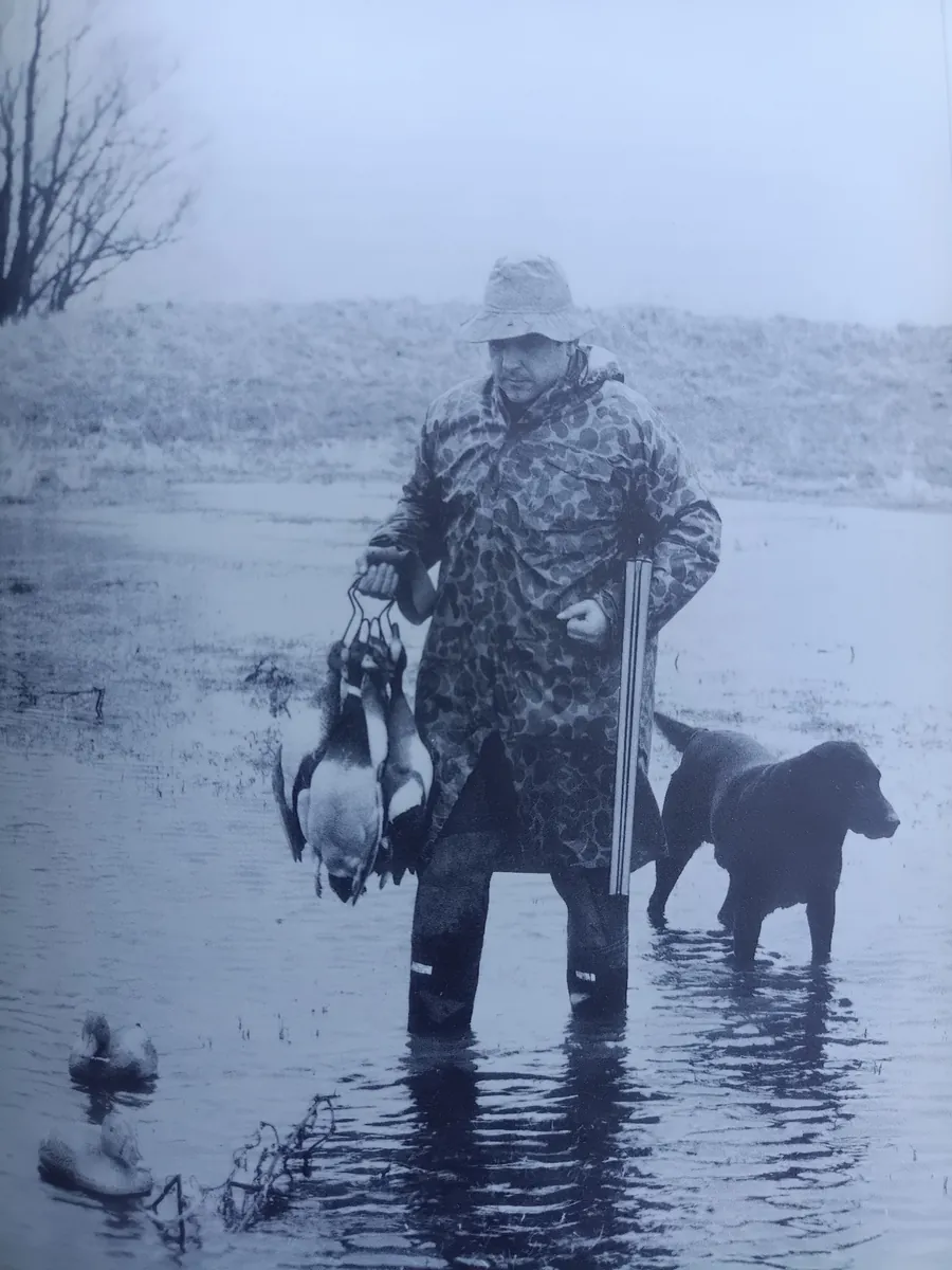 A wildfowler standing in water with a dog and birds carried at his side.